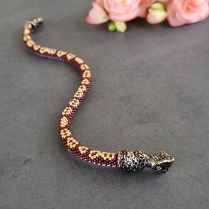 Puede incluir: Un collar con forma de serpiente hecho de cuentas en colores burdeos y dorado. El collar tiene un diseño detallado y texturizado y un cierre plateado. Hay flores rosas en el fondo.
