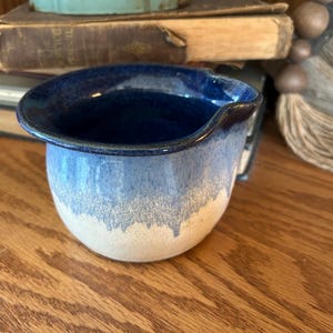 May include: A handmade ceramic pitcher with a dark blue rim and interior, transitioning to a light blue and white exterior. The pitcher has a rounded body and a small handle. It is sitting on a wooden surface.