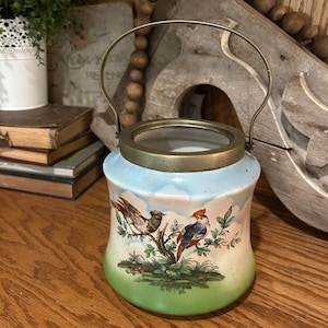 May include: A vintage ceramic jar with a metal handle and rim. The jar features a painted scene of birds perched on a branch, set against a sky and green landscape. The jar is cylindrical with a slight curve.