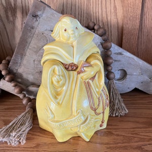 May include: Yellow ceramic cookie jar shaped like a monk holding a book and rosary beads. The jar has the text "Thou Shalt Not Steal" on the front.
