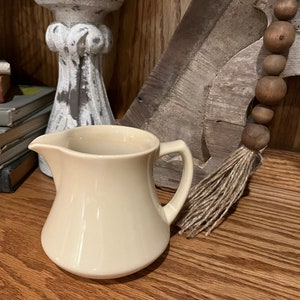 May include: A cream-colored ceramic creamer with a rounded body and a handle. The creamer is sitting on a wooden surface.