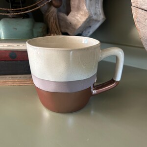 May include: A ceramic mug with a rounded base and a curved handle. The mug features a color-blocked design with a light beige top, a gray-purple middle section, and a brown base. The handle is white with a brown accent.