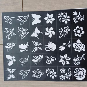 May include: A black stencil sheet featuring various white floral and butterfly designs. The sheet is divided into squares, each containing a different pattern. The designs include roses, butterflies, and other stylized flowers.