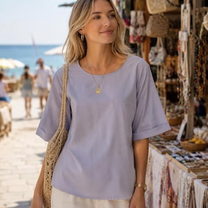 May include: A light lavender short-sleeved blouse with a round neckline and cuffed sleeves, paired with off-white trousers and a woven straw tote bag. The blouse features a simple, solid-coloured design with a relaxed fit and a straight hemline.