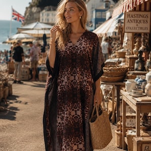 May include: A loose-fitting kaftan dress featuring a dark brown and black leopard print pattern with a V-neckline and wide sleeves. The garment has a long, flowing silhouette and is paired with a woven straw tote bag held at the side.