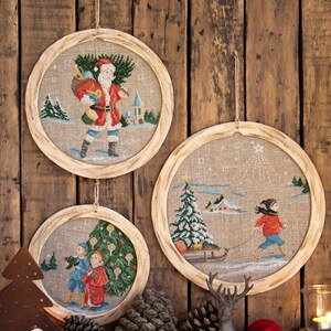 Puede incluir: Tres adornos redondos con marco de madera con escenas navideñas de invierno. Los adornos representan a Papá Noel, un niño tirando de un trineo y dos niños de pie junto a un árbol de Navidad. Los adornos están colgados en una pared de madera rústica.
