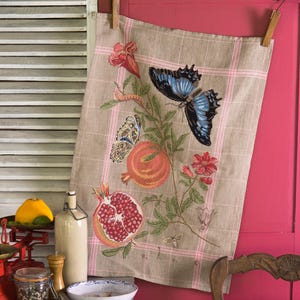 Puede incluir: Un paño de cocina beige con un estampado de cuadros rosas y bordados de mariposas, granadas y flores. El paño está colgado en una puerta roja. Una cesta con un paño a rayas rojas y blancas está junto a una silla de madera.