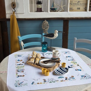 May include: A table set for a meal, with a white tablecloth featuring a seaside design. The table setting includes plates, cutlery, napkins, and a decorative serving tray. A seagull figurine and lighthouse decor are visible in the background.