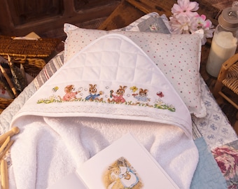 May include: White hooded towel with embroidered bunnies blowing bubbles. The towel is on a white and blue patterned bedspread with a book and wooden clothespins.