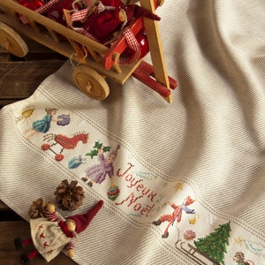 Könnte beinhalten: Ein weißes Leinen-Geschirrtuch mit einem Kreuzstich-Design, das eine Weihnachtsszene mit dem Text "Joyeux Noel" zeigt. Das Tuch ist gefaltet und liegt auf einer Holzoberfläche mit einem Spielzeugwagen, der mit roten Kugeln gefüllt ist.