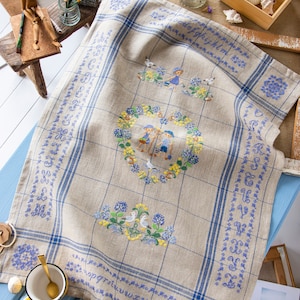 Puede incluir: Una toalla de té de lino beige con bordes bordados en azul y blanco. El centro presenta una corona en forma de corazón con dos niños tomados de la mano, rodeada de flores y pájaros. El borde está bordado con el alfabeto en azul.