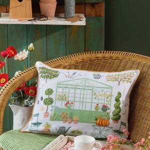 Peut inclure: Un coussin blanc avec un motif brodé d'une serre avec des plantes et des outils de jardinage. Le coussin est placé sur un fauteuil en osier avec un plateau contenant une tasse de thé et une assiette de biscuits.