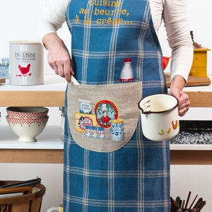 May include: A blue and white plaid apron with a large pocket featuring a cross-stitch design of kitchen items and the text "Cuisine au beurre, à la crème..." in gold thread.