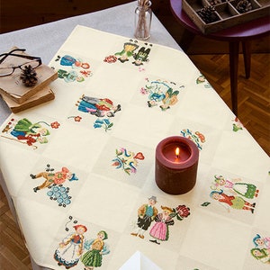 Könnte beinhalten: Eine weiße Tischdecke mit einem Kreuzstichmuster, das Paare in traditioneller Kleidung zeigt. Die Tischdecke ist mit floralen Motiven verziert und hat eine rote Kerze in der Mitte.