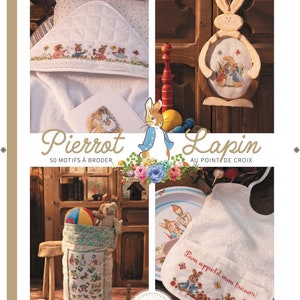 Peut inclure: Une couverture de livre représentant un lapin blanc vêtu de bleu et portant une couronne de fleurs roses. Le titre du livre est "Pierrot Lapin" avec le texte "50 Motifs à Broder" et "Au Point de Croix". La couverture présente également une serviette blanche avec une broderie de lapin, un lapin en bois avec une broderie de lapin et un bavoir blanc avec une broderie de lapin.