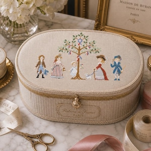 May include: An oval beige box with a cross-stitch design of a family under a tree. The box has gold trim and a decorative clasp. The text "Rubans de France PARIS" is visible. Ribbons, scissors, and a ribbon spool are also present.