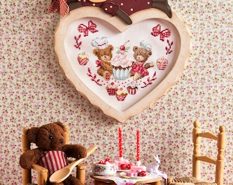 Könnte beinhalten: Ein herzförmiger Holzrahmen mit einem Kreuzstich-Design von drei Teddybären, die sich einen Cupcake teilen. Der Rahmen hängt an einer floralen Tapete. Im Vordergrund stehen zwei Holzstühle und ein Tisch mit einem Puppen-Tee-Set.