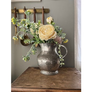 Può includere: Un'anfora d'argento con finitura martellata contiene un bouquet di fiori di seta color crema con fogliame verde.