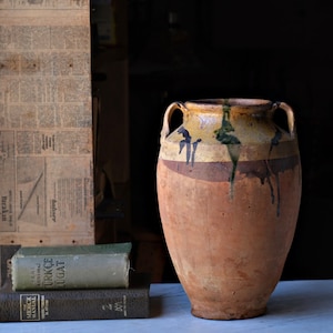 May include: A large, brown, earthenware jar with a wide mouth and two handles. The jar has a yellow and brown glaze with black and green accents. The jar is sitting on a white surface.