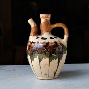 May include: A brown and white ceramic pitcher with a green and brown glaze. The pitcher has a wide mouth and a handle.