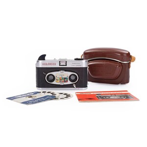 May include: A silver View-Master camera with a black body and a brown leather case. The camera has a red and white label that reads "View-Master" and "Redenstock Trion 1:3.5/35mm". The camera is accompanied by a set of 3 reels.