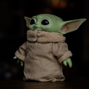 May include: A green-skinned figure with large black eyes and pointed ears, wearing a tan coat with a fuzzy, cream-coloured collar and cuffs. The figure is a popular character from a science fiction series.
