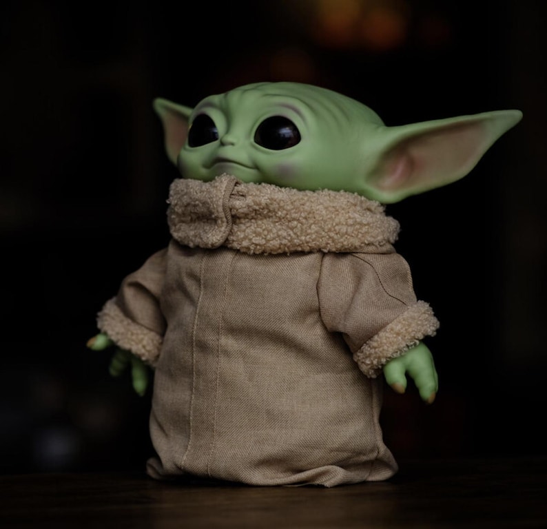 May include: A green-skinned figure with large black eyes and pointed ears, wearing a tan coat with a fuzzy, cream-coloured collar and cuffs. The figure is a popular character from a science fiction series.