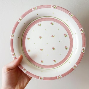 Puede incluir: Un plato de cerámica blanco con un diseño floral rosa. El plato presenta un borde rosa con pequeños detalles florales y un anillo interior verde. Pequeñas flores rosas y puntos verdes están dispersos por el centro blanco.