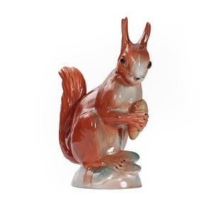 Puede incluir: Una figurita de porcelana de una ardilla roja sosteniendo una nuez en sus patas. La ardilla está sentada sobre una base con hojas verdes y blancas.