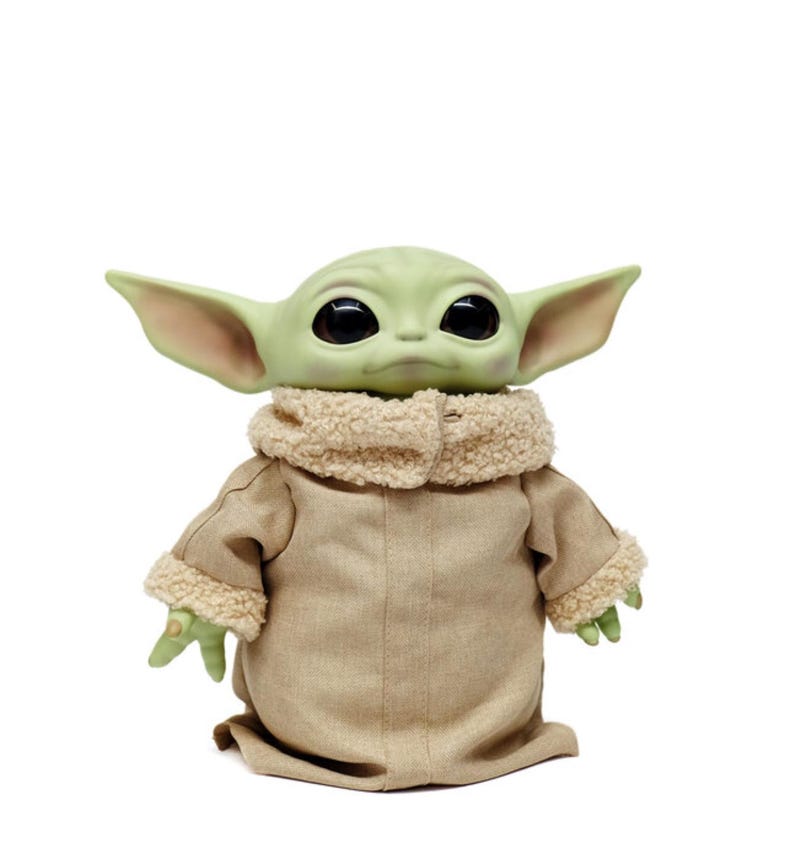 May include: A plush doll of a character with large black eyes, green skin, and large ears. The doll is wearing a tan coat with a fluffy, cream-coloured collar and cuffs. The doll is standing against a white background.