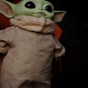 May include: A green-skinned doll with large black eyes and pointed ears. The doll is dressed in a tan coat with a fluffy, cream-coloured collar. The doll is a character from a popular science fiction series.
