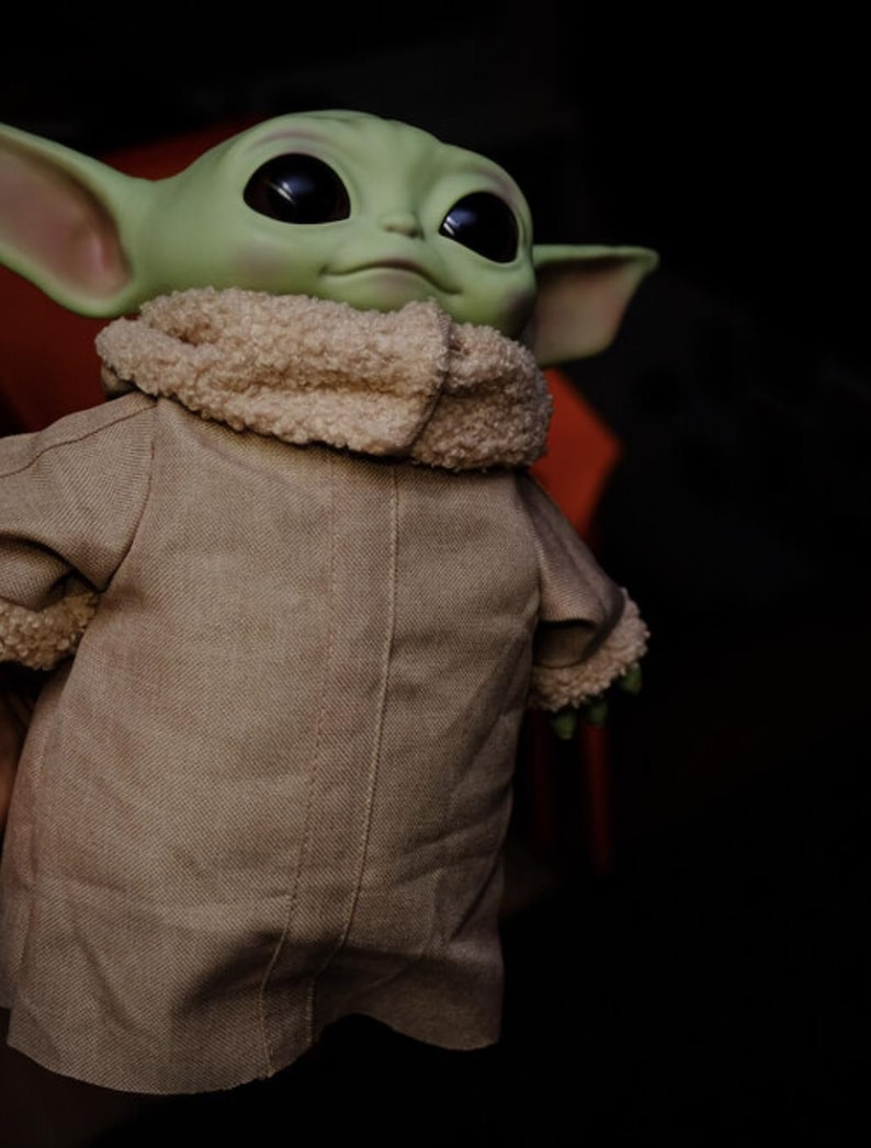 May include: A green-skinned doll with large black eyes and pointed ears. The doll is dressed in a tan coat with a fluffy, cream-coloured collar. The doll is a character from a popular science fiction series.