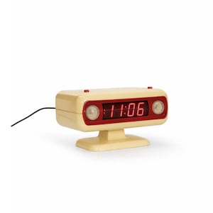 May include: A retro-style digital alarm clock in cream and red. The clock displays the time 11:06 in red numerals. It has a rectangular face with two circular lights and a small base. A black power cord extends from the back.