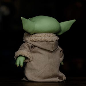 May include: A soft toy of a green-skinned character with large ears, wearing a tan robe with a fluffy collar and cuffs. The figure is seen from the back, against a dark background.