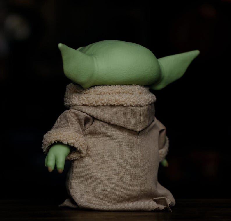 May include: A soft toy of a green-skinned character with large ears, wearing a tan robe with a fluffy collar and cuffs. The figure is seen from the back, against a dark background.
