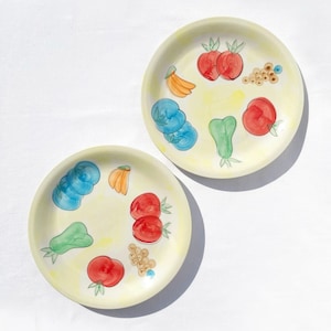 May include: Two white ceramic plates with yellow backgrounds and colourful fruit illustrations. The plates feature painted images of apples, bananas, plums, and grapes.