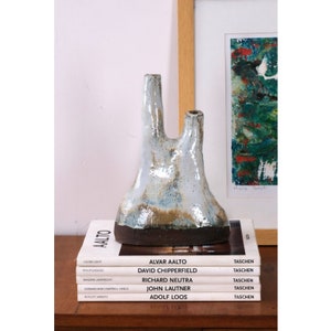 May include: A white ceramic vase with a unique, organic shape and a brown base. The vase has two openings at the top, making it ideal for displaying a variety of flowers or greenery. The vase is sitting on top of a stack of books with the titles "Alvar Aalto", "David Chipperfield", "Richard Neutra", "John Lautner", and "Adolf Loos".
