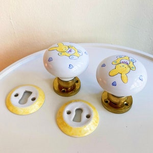 以下が含まれることがあります： 黄色と青のウサギのデザインが施された白いセラミック製のドアノブが2つ。ノブは金色のベースで、白い表面の上に置かれています。