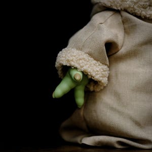 May include: A tan fabric garment with a cream-coloured, fuzzy trim. A green hand with three fingers and a thumb emerges from the garment. The background is black.