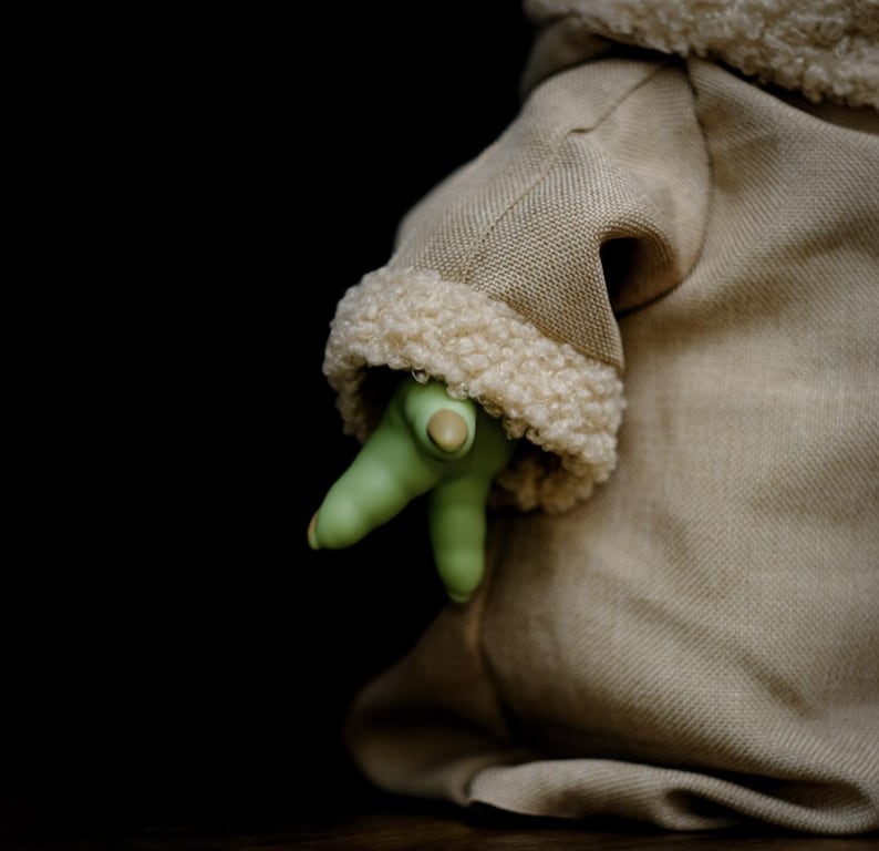May include: A tan fabric garment with a cream-coloured, fuzzy trim. A green hand with three fingers and a thumb emerges from the garment. The background is black.
