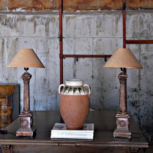May include: Two lamps with beige lampshades and brown bases sit on a dark wood table with a brown ceramic vase and a stack of books. The lamps are on a table in front of a concrete wall with rusty pipes.