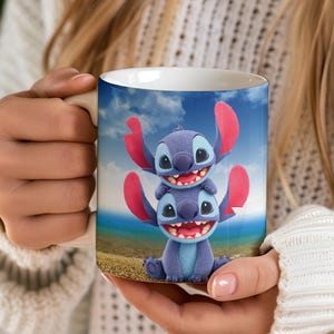 Puede incluir: Taza de cer&aacute;mica blanca con un dise&ntilde;o de personaje de dibujos animados en azul y rosa. El dise&ntilde;o muestra dos personajes apilados con orejas grandes y caras sonrientes sobre un fondo de cielo azul y playa. La taza es sostenida por una persona.
