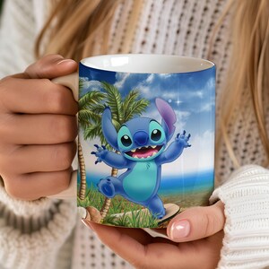 Puede incluir: Una taza de cer&aacute;mica blanca con una ilustraci&oacute;n vibrante de Stitch de Lilo & Stitch. El dise&ntilde;o muestra a Stitch en una playa con palmeras y un cielo azul. La taza es sostenida por una persona que lleva un su&eacute;ter blanco.