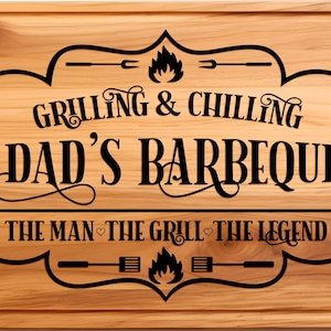 May include: A wooden cutting board with a black engraved design that says "Grilling & Chilling Dad's Barbeque The Man - The Grill - The Legend".