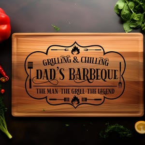 May include: A wooden cutting board with the text "Grilling & Chilling Dad's Barbeque! The Man - The Grill - The Legend" engraved on it. The board is surrounded by various vegetables and herbs.