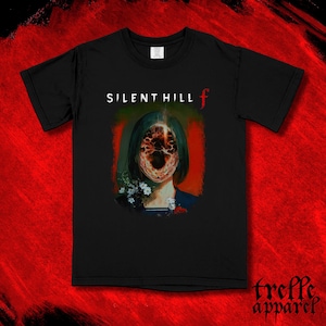 以下が含まれることがあります： 白と赤で「SILENT HILL f」の文字が入った黒いTシャツ。グラフィックは、顔が歪んだ人物、花、緑と赤の配色を描いています。Tシャツは赤い背景にあります。