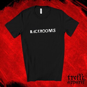 May include: A black short-sleeved t-shirt featuring the word BACKROOMS printed in white capital letters across the chest. The garment has a crew neckline and a standard fit, with a white brand label visible at the inside of the collar.
