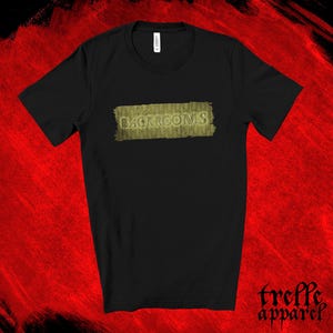 May include: A black short-sleeved t-shirt featuring a rectangular graphic on the chest. The graphic displays the word BACKROOMS in a stylized font over a textured, mustard-yellow background that resembles aged wallpaper with a repeating pattern.