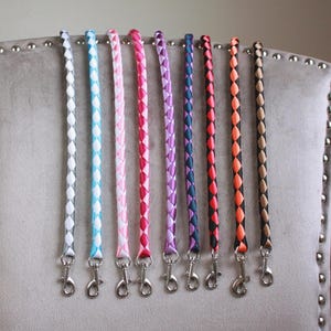 May include: Nine braided straps in a range of colours, including white, blue, pink, purple, navy, orange, and tan. Each strap has a silver clasp at the bottom. The straps are displayed against a grey upholstered chair.