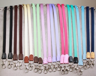 May include: A collection of twenty-two flat lanyards in various solid colors including black, brown, cream, green, lavender, pink, turquoise, and navy blue. Each lanyard features a silver-toned metal swivel lobster clasp at the bottom end.